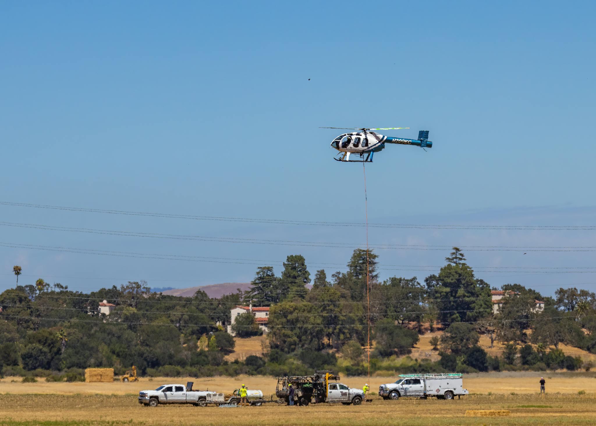 Helicopter in action over a rural field with trucks and team on a sunny day.